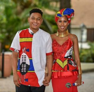 Swati bride in red Emahiya spaghetti strap peplum top with red skirt and headwrap. Accessorized with Ndebele gold choker and bangles. Swati groom in Emahiya print shirt.